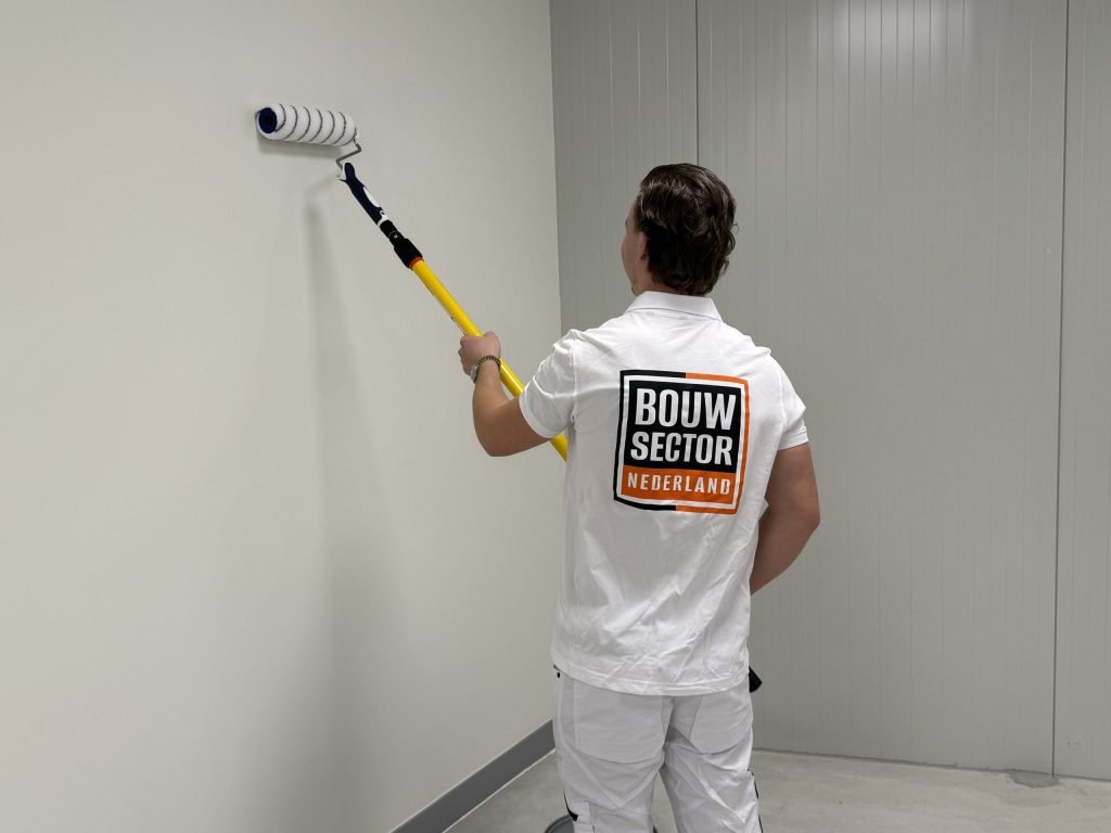 Schilder met logo van Bouwsector Nederland rolt een kale lichte wand in een lege ruimte