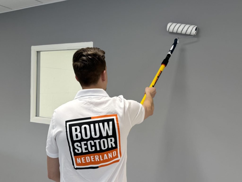Schilder in wit poloshirt met logo van Bouwsector Nederland rolt een grijze wand naast een wit raamkozijn