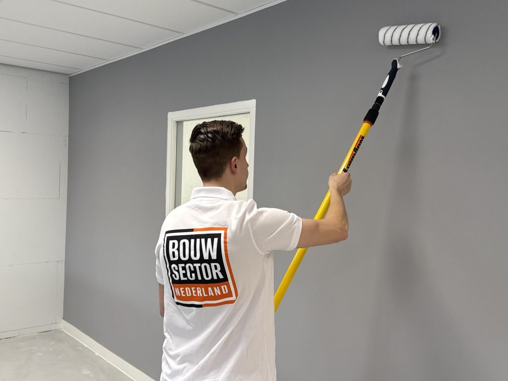 Schilder in wit werkshirt rolt grijze muurverf op een wand naast een raam met wit kozijn
