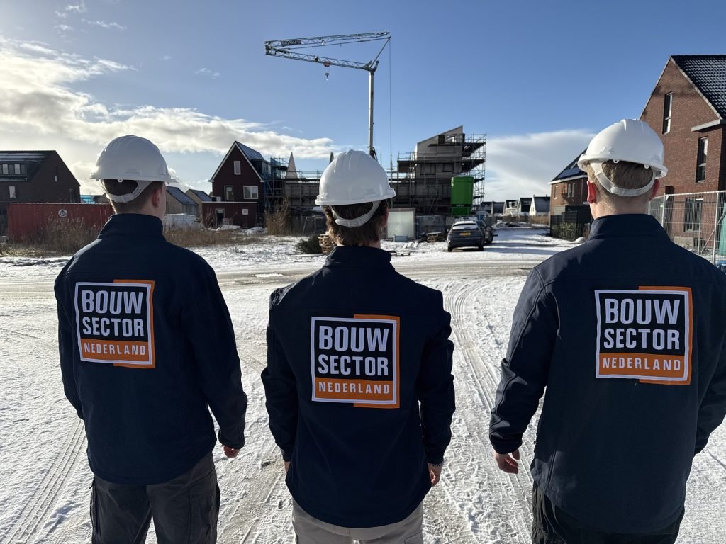 Drie vakmensen in donkerblauwe jassen met logo en witte helm kijken uit over nieuwbouw in de sneeuw