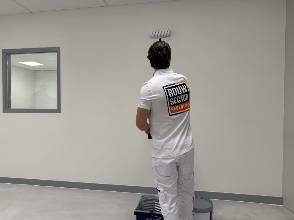 Schilder van Bouwsector Nederland zet met de roller een witte wand in de verf, met de verfbak op de vloer
